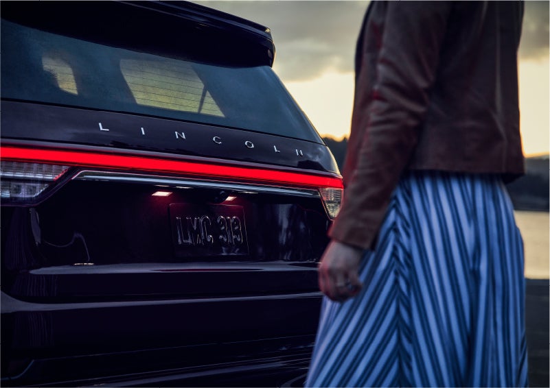 A person is shown near the rear of a 2023 Lincoln Aviator® SUV as the Lincoln Embrace illuminates the rear lights | Dave Sinclair Lincoln St. Peters in Saint Peters MO
